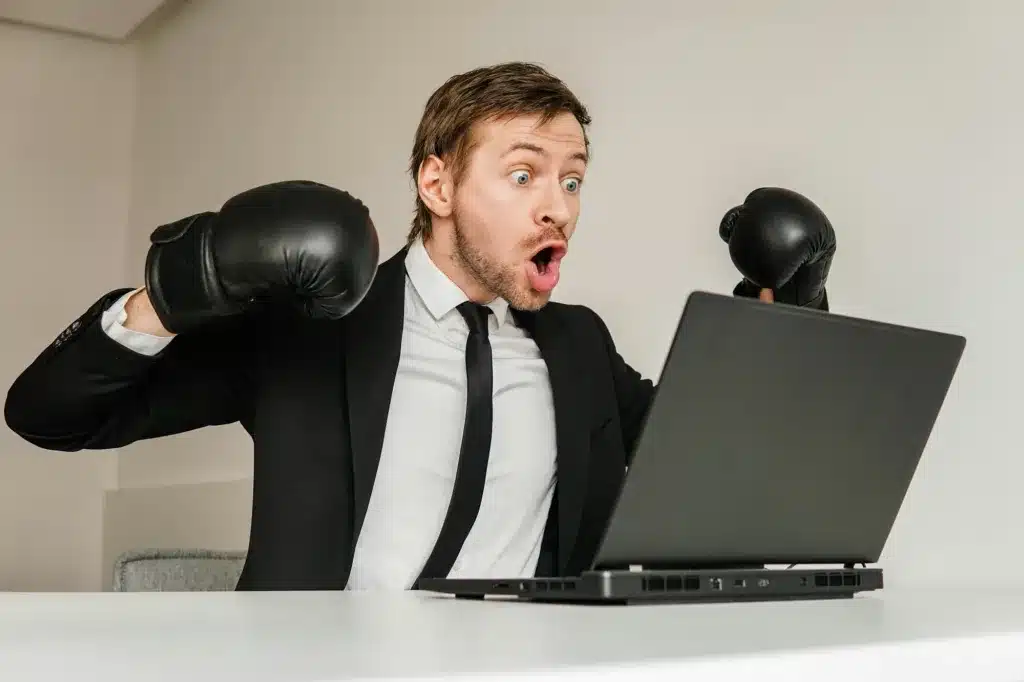 Young angry businessman in black suit wearing boxing gloves using laptop.
