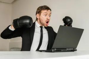 Young angry businessman in black suit wearing boxing gloves using laptop.