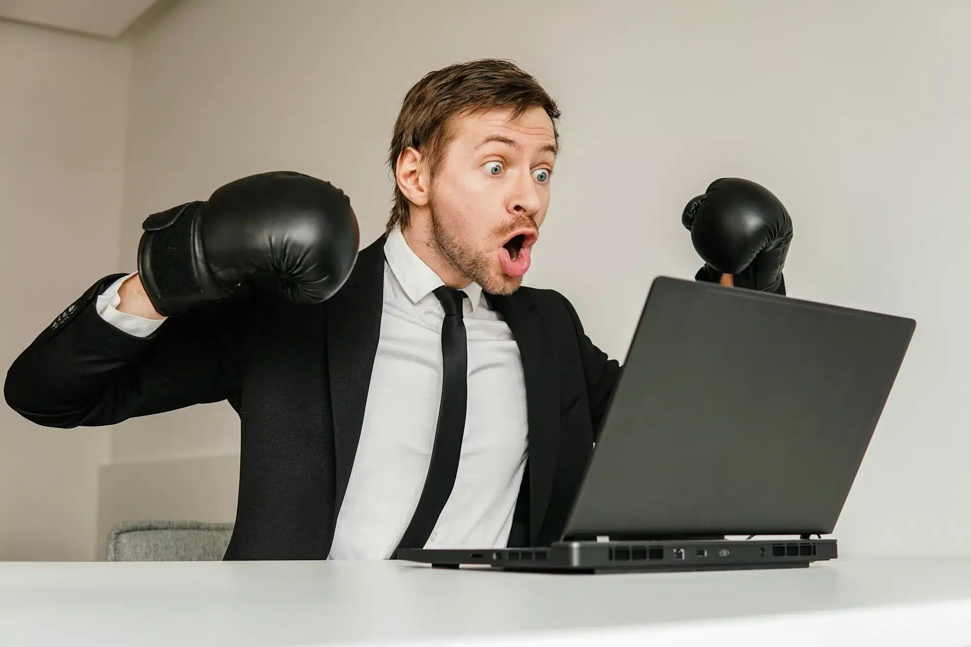 Young angry businessman in black suit wearing boxing gloves using laptop.